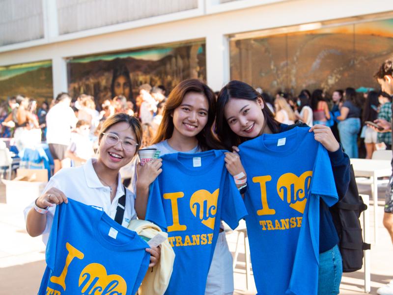 Three students holding shirts that say "I love UCLA transfers" at an event.